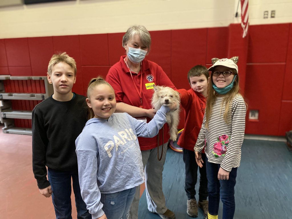 A group of children stand near an adult holding a therapy dog