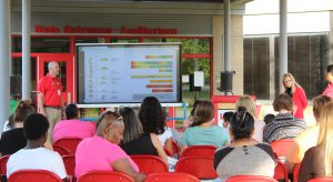 Dr. Sullivan speaks in front of a screen showing curricular updates as a dozen and a half people watch while sitting in red chairs