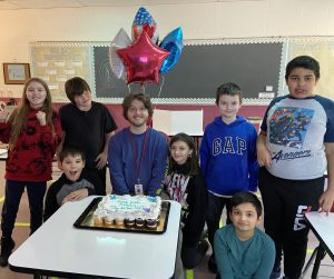Students and a veteran stand near a desk with a cake