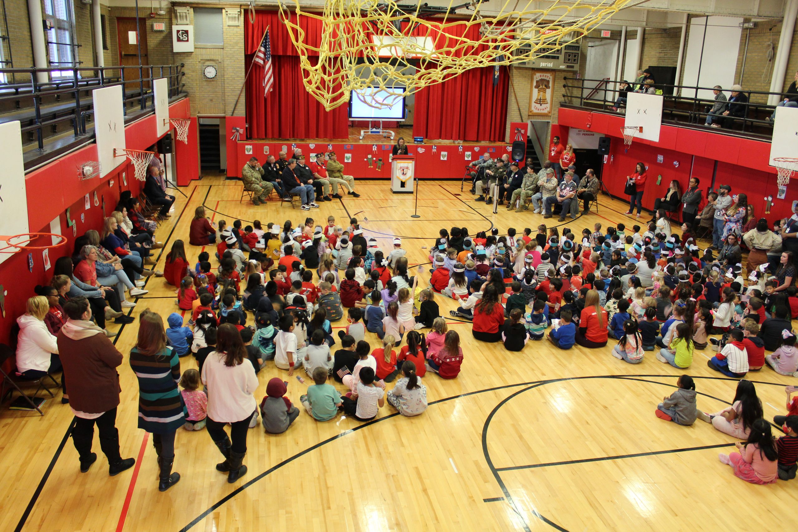 A person stands at a podium with veterans seated on either side as children sit on the floor in front of them