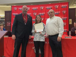 A woman holding a certificates stands between two men with the Liberty CSD banner behind them.