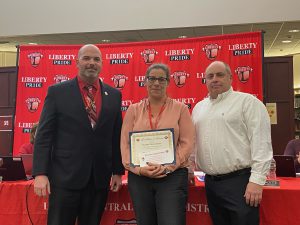 A woman holding a certificates stands between two men with the Liberty CSD banner behind them.