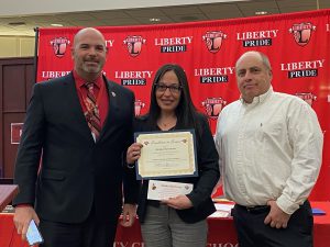 A woman holding a certificates stands between two men with the Liberty CSD banner behind them.