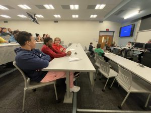Students sit at tables on risers facing forward