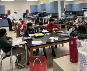 A chef prepares a meal as students sit at tables