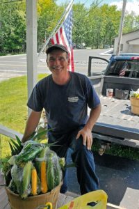 A man with a bushel of corn in front of him smiles while standing behind a pickup truck