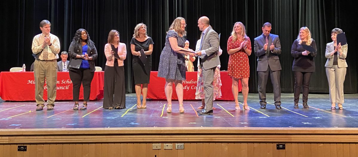 A woman at center stage is handed an award by a man as others standing in a line clap