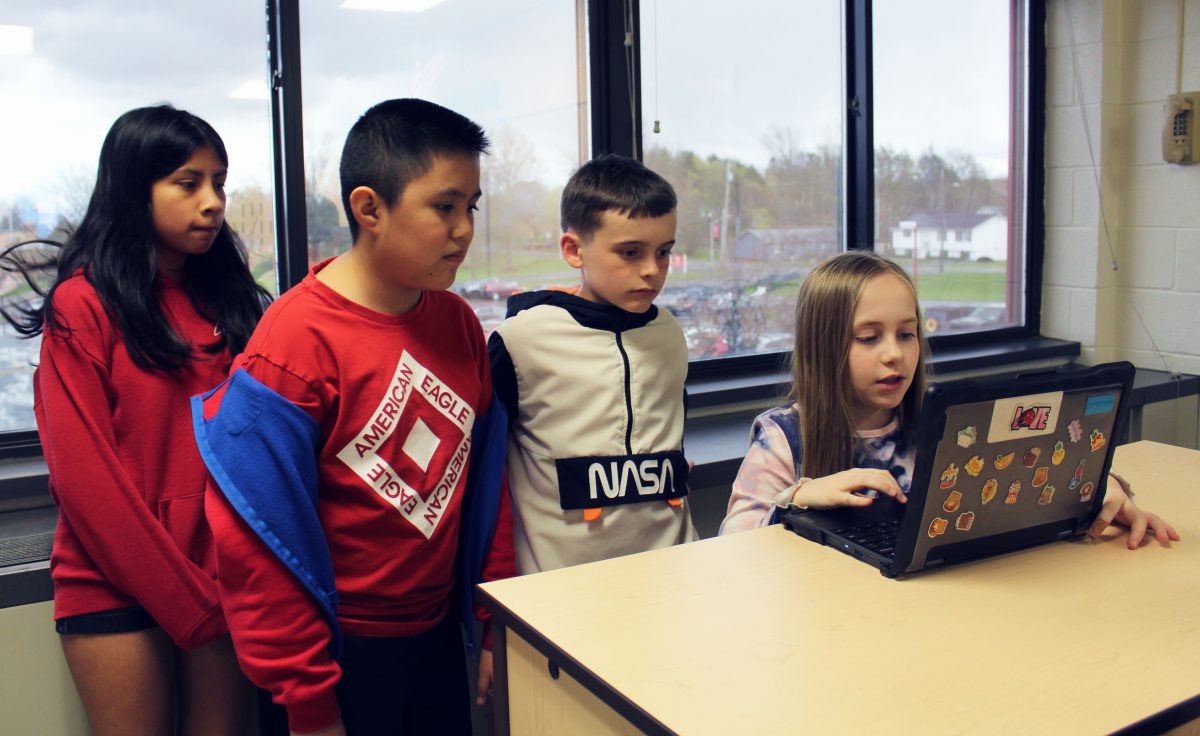 Four students stand behind a computer as they record a video