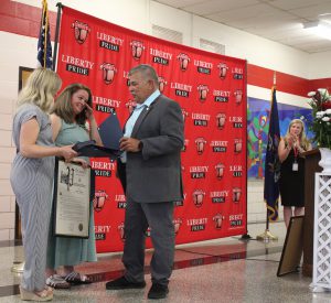 A man hands a proclamation to two women at left in front of a Liberty Pride banner, as a woman watches at right.