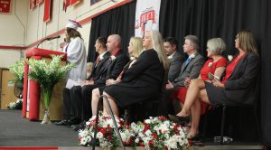 A student in a cap and gown speaks at a podium on a stage as adults sit in two rows behind her