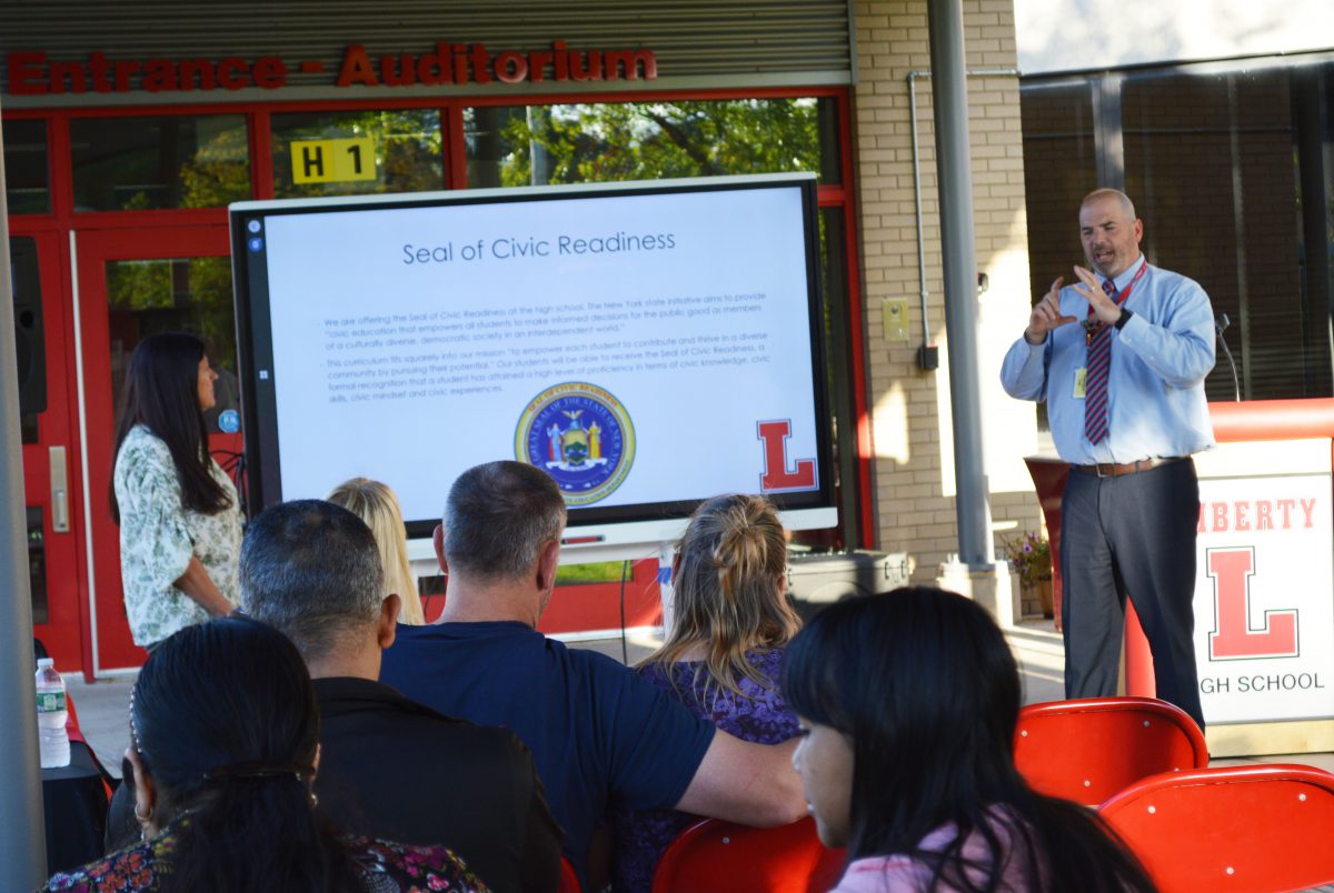 A man gestures at right as a woman stands at left with a large screen displaying a page of a presentation as a crowd seated in red chairs watch.