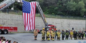 An American flag is suspended between two ladder trucks as people in firefighting gear stand near.