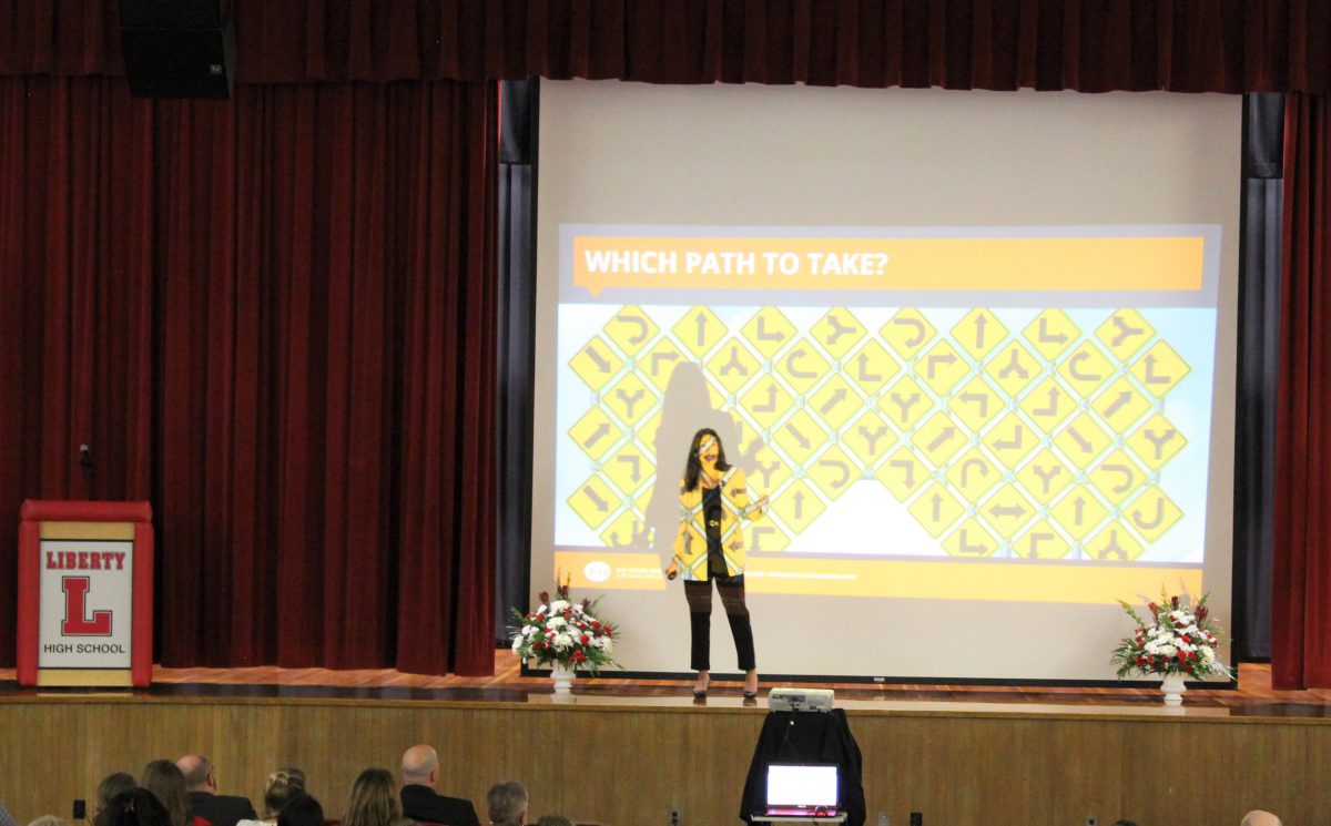 A woman stands on a stage with various directional signs with the words "Which Path to Take?" above shown on her and a screen behind her.