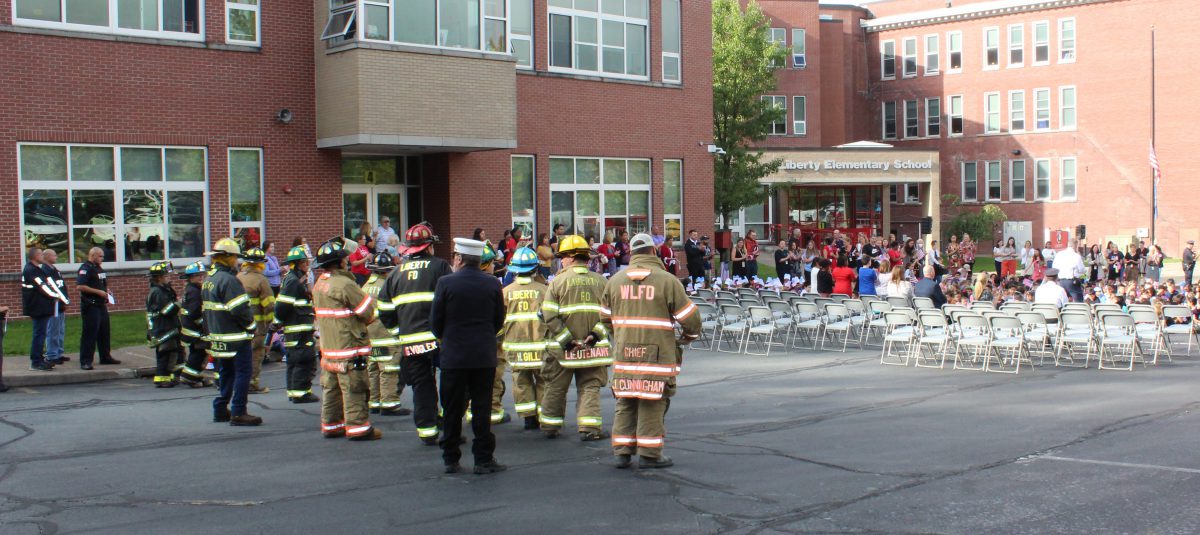 People in firefighting gear and first responder uniforms stand as students watch a ceremony