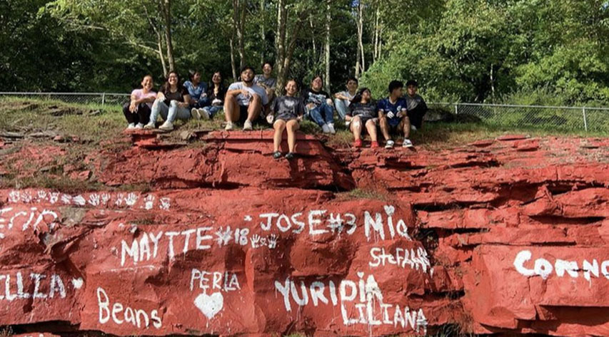 Students sit on top of a rock ledge painted red with white names and icons painted on it.