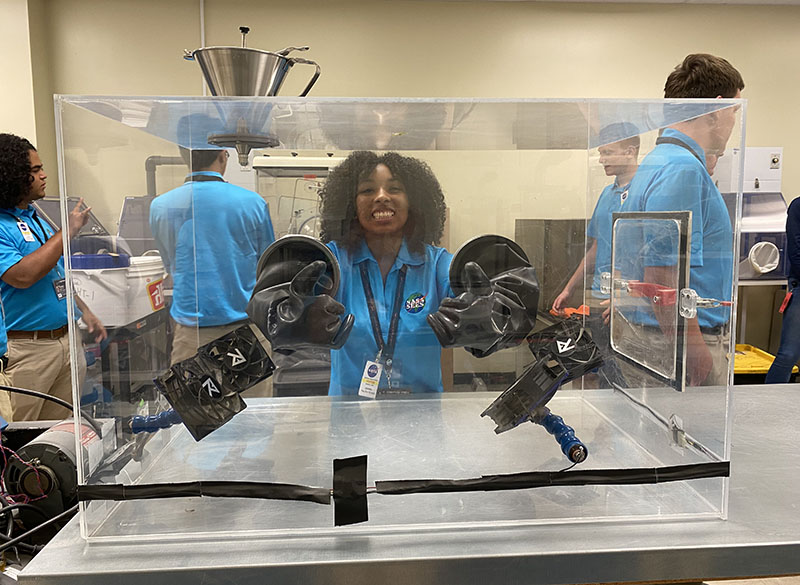 A student in a blue collard shirt with NASA on the breast hands into black arm-length gloves sealed to a clear box gives a thumbs up