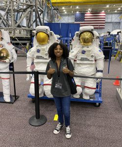 A student in a gray shirt gives a thumbs up in front of space suits on display.