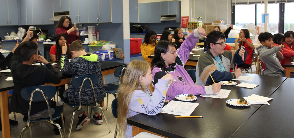 Students seated at tables give thumbs ups and thumbs down after a taste test.