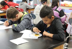 A student looks at a bite of food on a plate as another student writes on a piece of paper