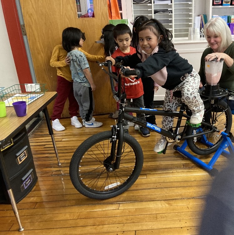 A student rides a bike with a blender attached at the back as a teacher holds the top of the blender and other students wait in line