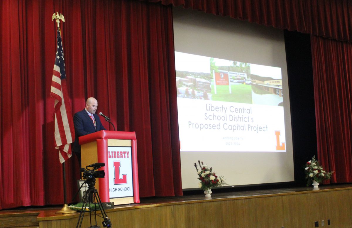 A man stands at a podium with an L on the front and flag beside him, with a screen featuring a slide that reads Liberty Central School District's proposed capital project
