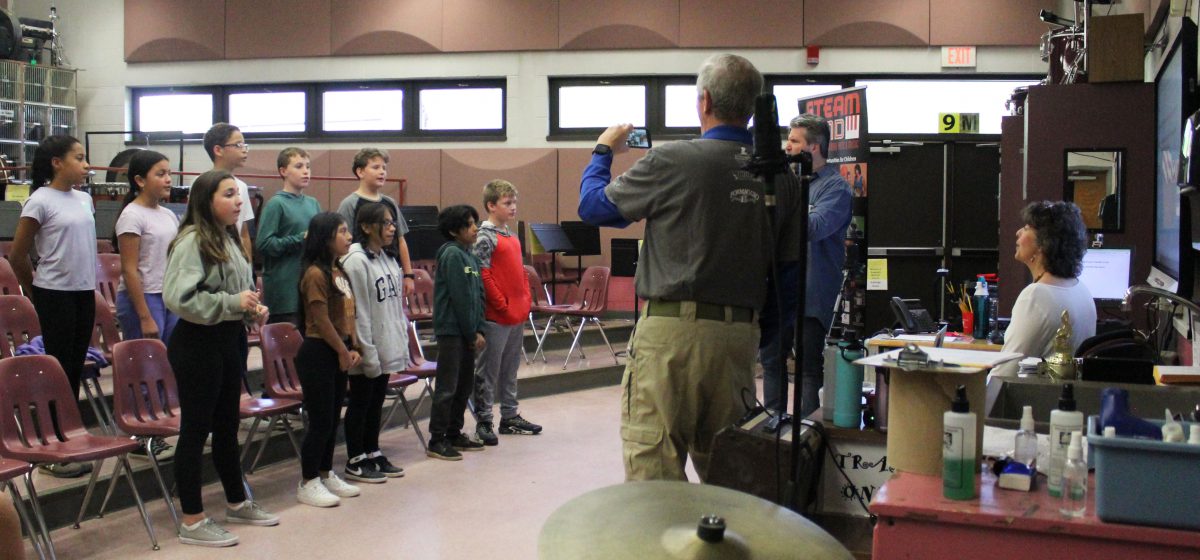 Students sing while standing on risers as a man videotapes with his phone and two people direct them