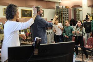 A man with his right pointer finger up and a woman holding her right palm out direct students as they sing