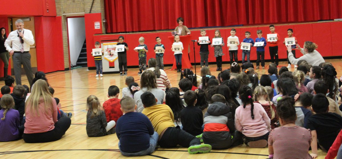 Students stand in a line displaying certificates as a person stands behind them and to the left and other students watch while sitting on the gym floor