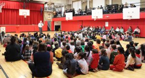 A man speaks into a microphone in a gym as people sit on the floor and other watch from seats above.