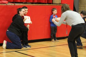 A woman kneels on the floor with her arm around a child holding a certificate as another person takes a picture.  Another child holding a certificate is in the background.