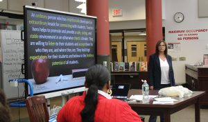 A woman stands to the right of screen with a display about educational superheroes as a teacher sits in the foreground