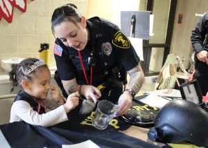 A police officer shows a young student how to dust for fingerprints on a glass.