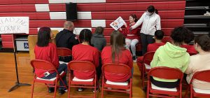 A student reads a book to an audience seated in chairs as another student holds a microphone for her