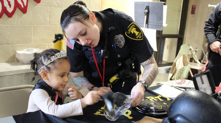 A young student in a tiara uses a brush to dust for fingerprints on a glass as a police officer helps.