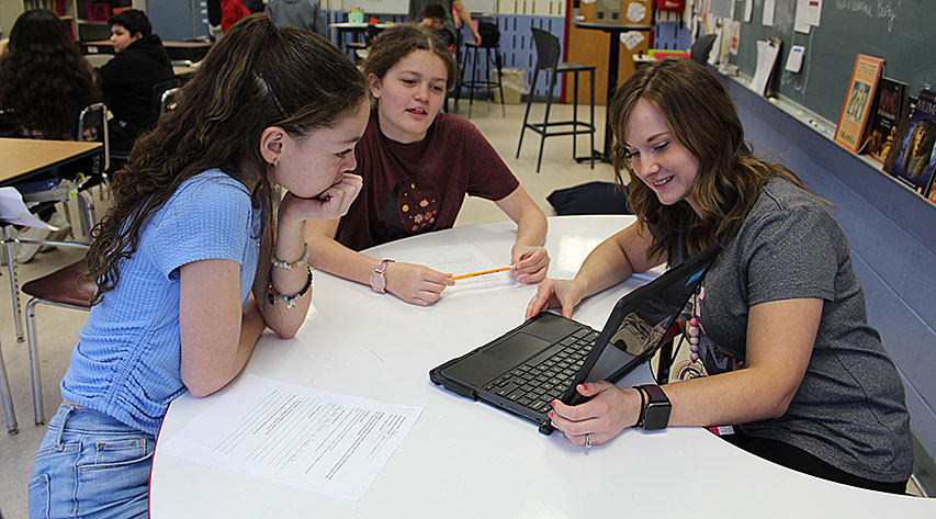 A teacher points out something on her laptop screen to two students seated at a table