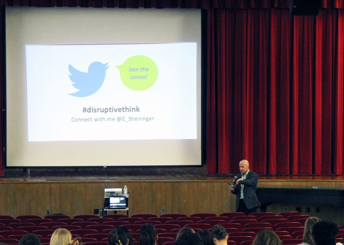 A man speaks with a screen behind him at the front of an auditorium