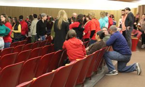 a person kneels behind a row of seats with others seated in them as the people speak in a group.