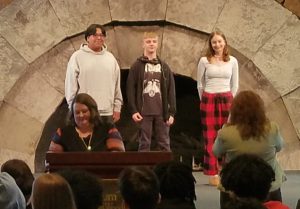 Three student stand on a stage as an adult speaks into a microphone.