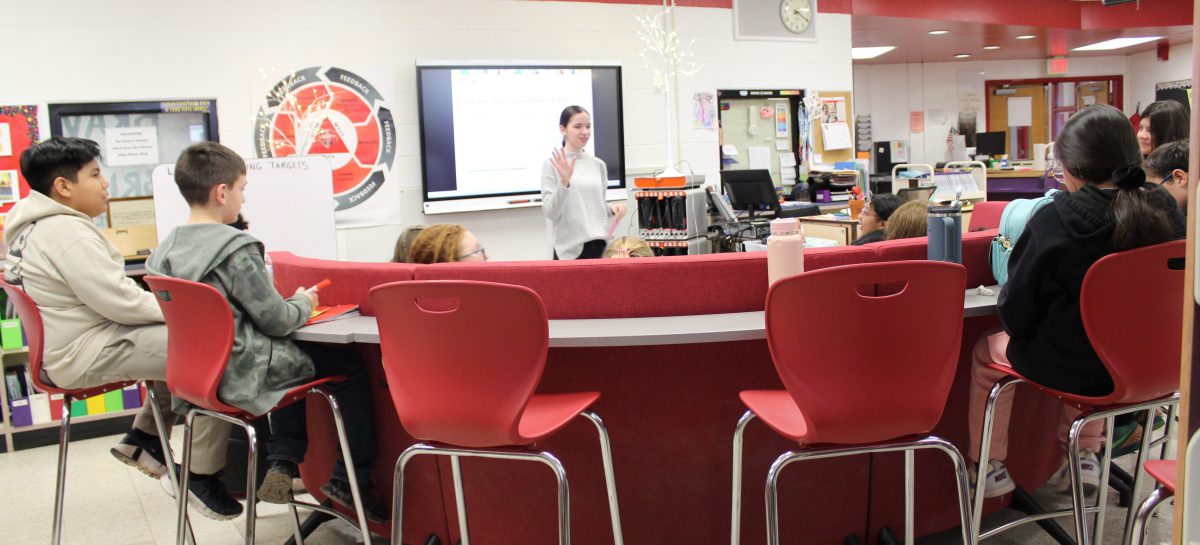 Students sitting around a curved desk listen to an adult speak in front of a screen.