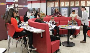 Students seated at a curved table look at pamphlets as an adult stands at the front of the room