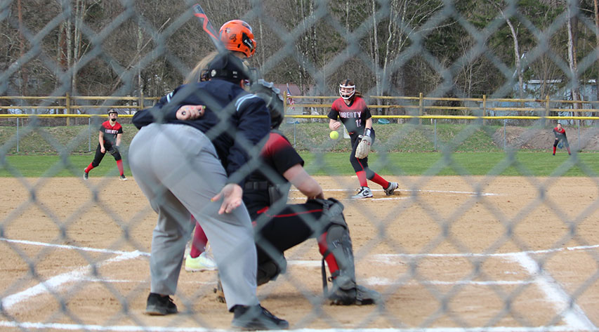 A softball pitcher throws to the catcher as a batter stands and an up watches, as seen through a chain link fence.