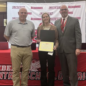 A woman holds a certificate and a a piece of paper with a pin on it flanked by two men.