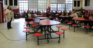A person speaks in front of a large screen as a crowd watcher her seated at round tables in a cafeteria