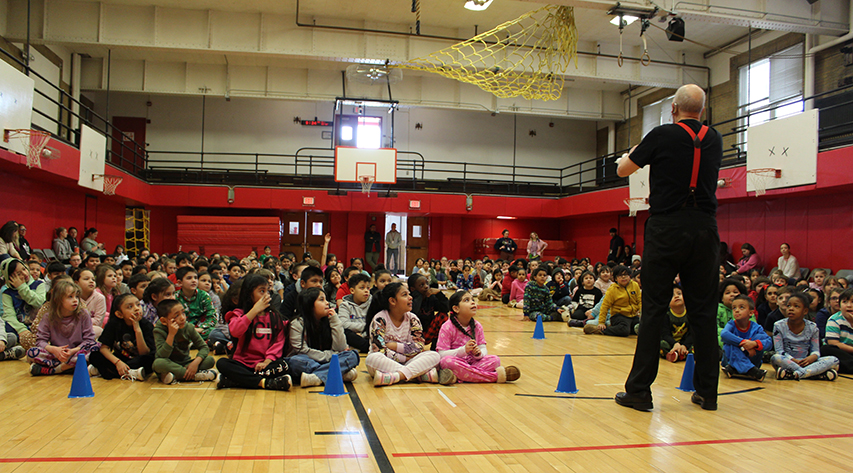 A man performs a magic trick in front of elementary students seated on a gym floor