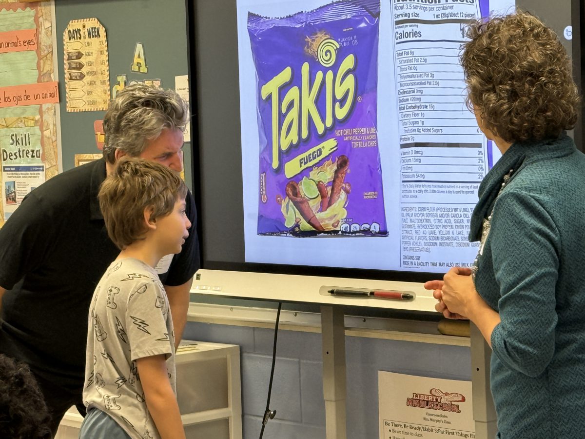 A student looks at a screen with a Takis bag and an ingredient list on it as two adults watch.