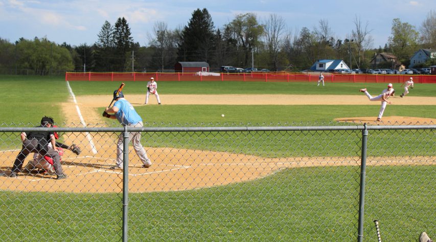 A pitcher throws a ball as his teammates go in motion as the batter watches the ball come toward him and the umpire and catcher watch from behind the plate.