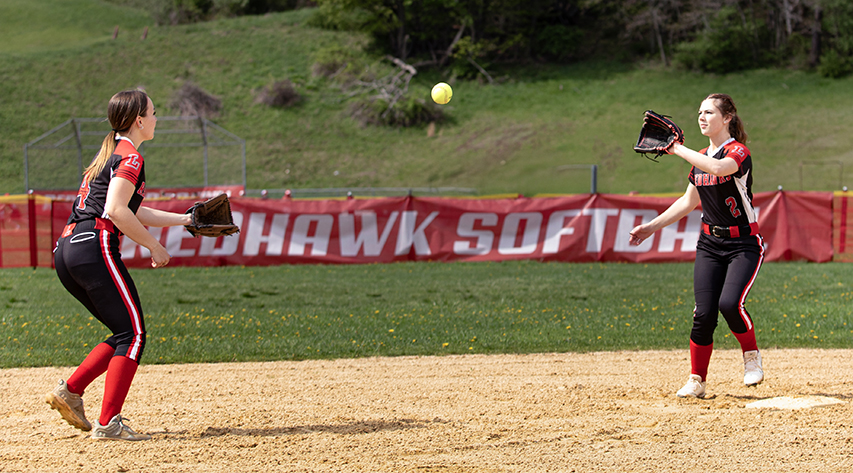 Two softball players throw the ball to each other in front of the Redhawk Softball sign on the diamond