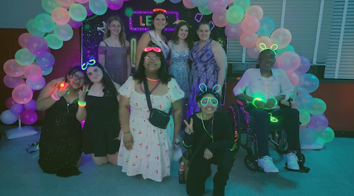 Students in formal attire and neon accessories pose for a photo under a neon balloon arch