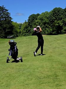 A golfer swings with his bag behind and the left to him