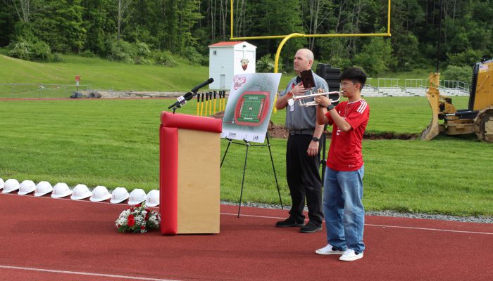 A student plays the trumpet as a man stand behind a podium with his hand on his heart